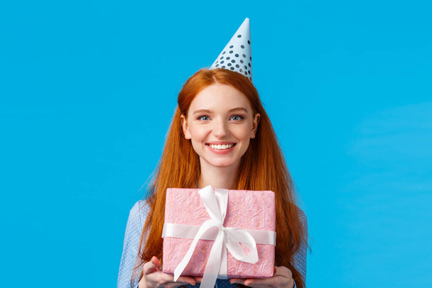 Waist-up portrait cheerful happy, pretty redhead, ginger girl celebrating birthday, receive cute pink wrapped present, wearing b-day cap and smiling, having fun at party, blue background - Photo, Image