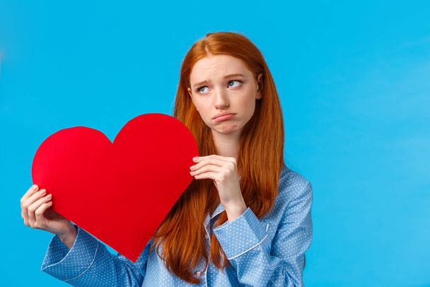 Gloomy girl was rejected on valentines day. Uneasy upset redhead female in nightwear, holding heart card and sobbing, look away upset, feeling depressed, standing blue background - Foto, Imagem