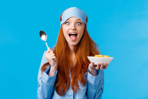 Femme caucasienne heureuse, excitée et accablée, mangeant des céréales le matin, tenant une cuillère, souriant, amusée et étonnée, regardant la caméra, portant un masque de sommeil, un pyjama, debout fond bleu - Photo, image