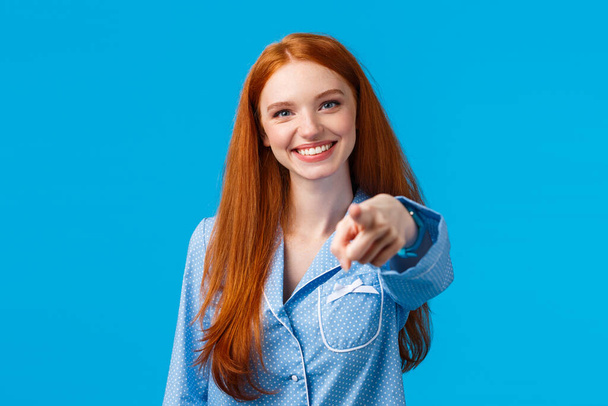 We need you. Cheerful and confident redhead girl making assured choice, pointing finger camera and smiling as decide, picking from variants, talking to person, standing in pyjama blue background - Zdjęcie, obraz