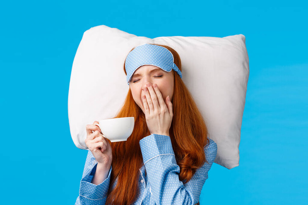 Sleepy cute redhead european girl with long ginger hair, wearing sleepwear, sleep mask close eyes and yawning, waking up morning, drinking coffee in bed, lying on pillow, blue background - Photo, Image