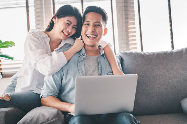 Πορτρέτο του ευτυχισμένου ασιατικού ζευγαριού στην αγάπη χαμογελώντας και γελώντας έχοντας μια καλή στιγμή μαζί στον καναπέ στο σπίτι με φορητό υπολογιστή - Φωτογραφία, εικόνα