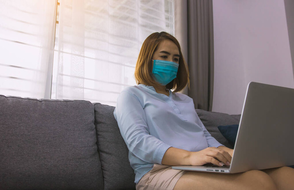 Aziatische vrouw werken vanuit huis aan een laptop computer tijdens een coronavirus epidemie door quarantaine en het dragen van medische maskers. - Foto, afbeelding