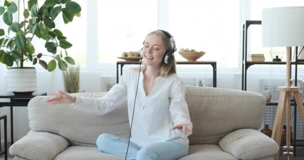 Joyful woman enjoying music using headphones on sofa - Materiał filmowy, wideo