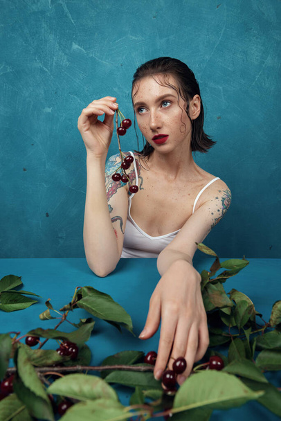 Dreamed female fashion model sitting at the table with cherry branch in front of the blue background  - Photo, Image
