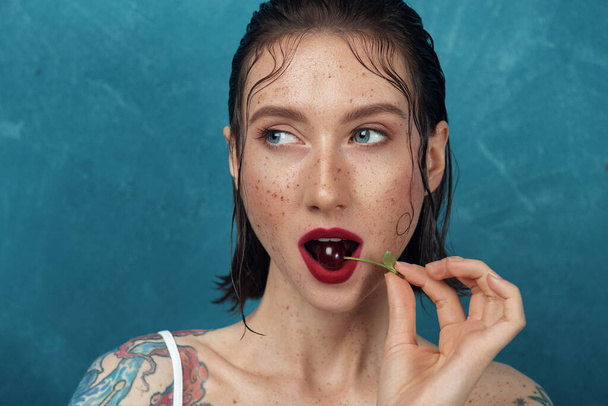 Nahaufnahme Porträt eines jungen Modells mit Kirschbeere. Sommersprossiges Mädchen beißt vor blauem Hintergrund in Kirsche. - Foto, Bild