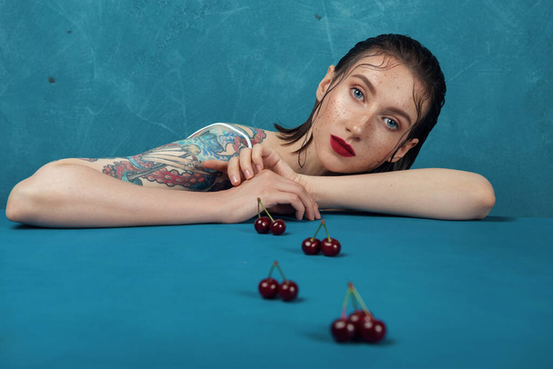 Young fashion model sitting at the table with cherry berries in front of the blue background - Photo, Image