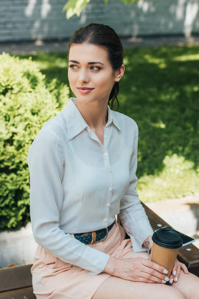 atractiva mujer de negocios sentada en el banco y sosteniendo la taza de papel
  - Foto, Imagen