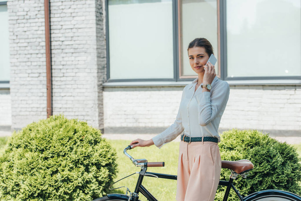 attractive businesswoman talking on smartphone and standing near bicycle  - Zdjęcie, obraz