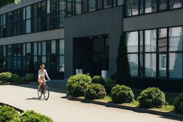 young businesswoman in glasses listening music in wireless headphones and riding bicycle near building  - 写真・画像