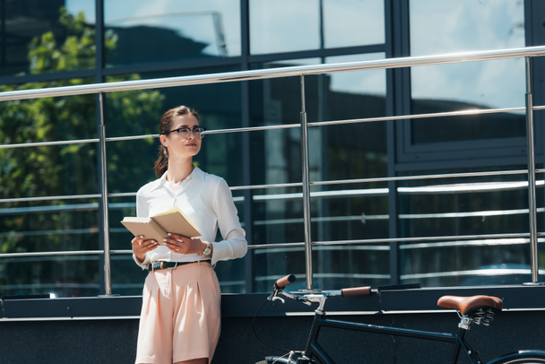 beautiful businesswoman in glasses holding book near bicycle and modern building  - Photo, Image