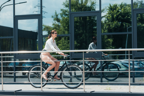 vrolijk jong zakenvrouw in bril rijden fiets in de buurt van gebouw  - Foto, afbeelding