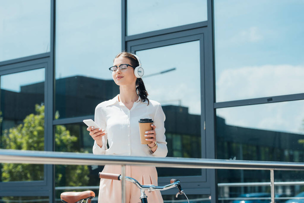 щаслива бізнес-леді в бездротових навушниках тримає смартфон і паперовий стаканчик біля будівлі
  - Фото, зображення