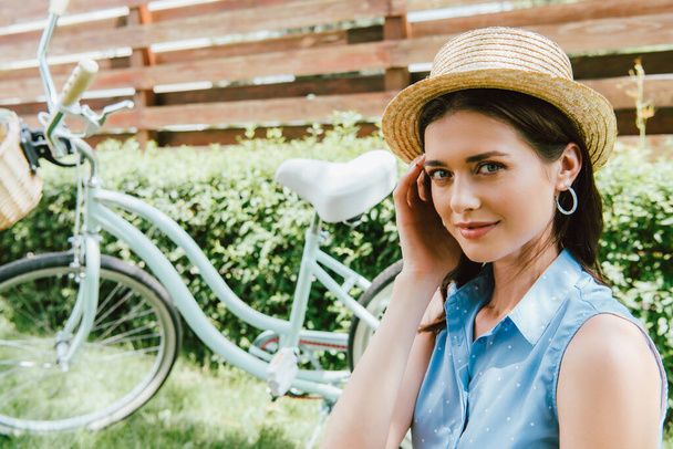 atrakcyjna kobieta dotykająca słomkowego kapelusza i uśmiechnięta w pobliżu roweru  - Zdjęcie, obraz