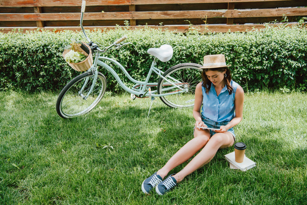 dziewczyna w słomkowym kapeluszu za pomocą tabletu cyfrowego i siedzi na trawie w pobliżu roweru, kubek papieru i książek - Zdjęcie, obraz