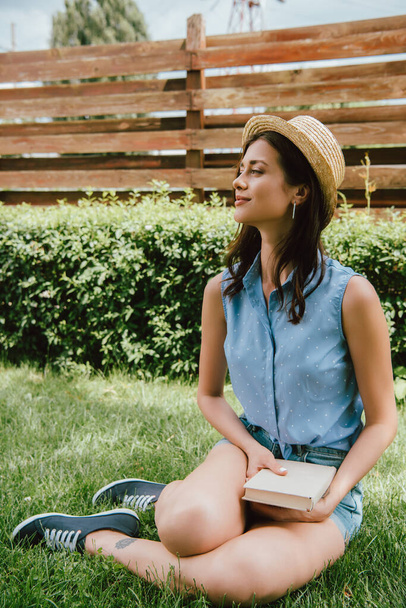 heureuse fille en paille chapeau tenant livre et assis sur l'herbe  - Photo, image