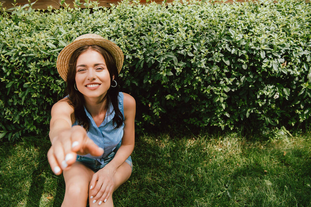 enfoque selectivo de la chica feliz en sombrero de paja con la mano extendida sentado cerca de arbusto
 - Foto, Imagen