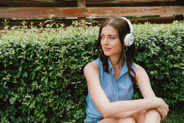 χαρούμενη νεαρή γυναίκα ακούγοντας μουσική σε ασύρματα ακουστικά κοντά σε θάμνους  - Φωτογραφία, εικόνα