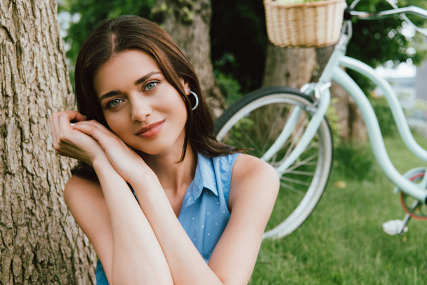 selective focus of attractive woman looking at camera near bicycle and tree trunk - Photo, Image