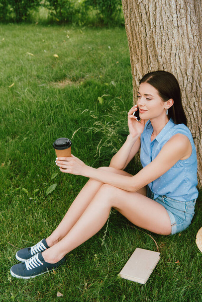 schöne Frau, die auf dem Smartphone spricht und Pappbecher in der Hand hält, während sie im Gras sitzt - Foto, Bild