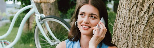 panoramic shot of happy woman talking on smartphone near bicycle  - Photo, Image