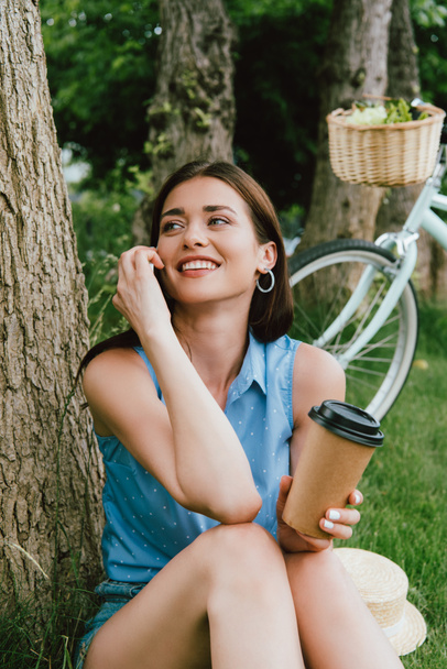 smiling woman talking on smartphone and holding paper cup while sitting near tree trunk - Φωτογραφία, εικόνα