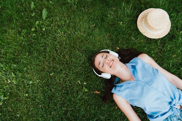 top view of cheerful woman lying on grass and listening music near straw hat  - Fotografie, Obrázek
