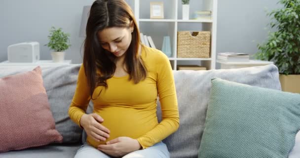 Portrait shot of the beautiful pregnant woman sitting on the sofa, caressing her belly and smiling to the camera in the nice room at home. Indoors - Filmmaterial, Video