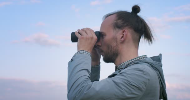 Side portrait of hipster bearded man looking into binoculars afar handheld camera slow motion. Authentic man with eastern hairstyle holding glasses outdoors copy text space. Travel adventure equipment - Footage, Video
