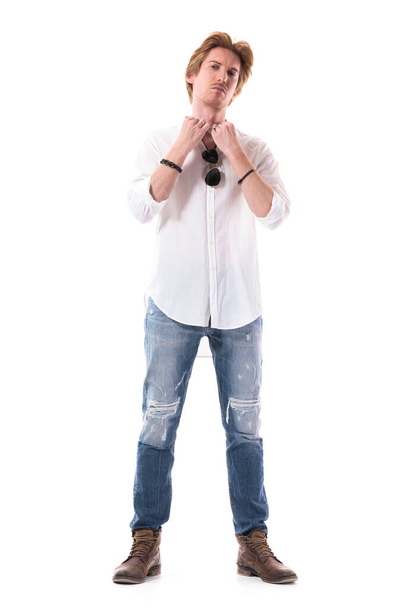 Σοβαρός νεαρός κομψός άντρας ντύνεται προσαρμόζοντας το γιακά του πουκάμισου κοιτάζοντας την κάμερα ως καθρέφτη. Πλήρες μήκος σώματος που απομονώνεται σε λευκό φόντο.  - Φωτογραφία, εικόνα