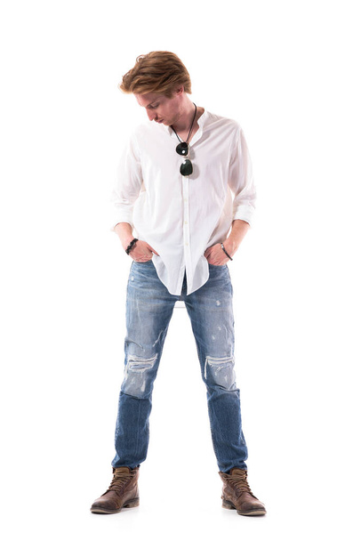 Красивый молодой человек покупает и примеряет новые порванные джинсы, глядя вниз. Полная длина тела изолирована на белом фоне
.  - Фото, изображение