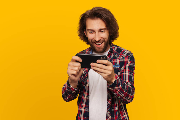 Joyful young man plays a game on a smartphone, a man with long hair and a shirt plays on a mobile phone, on a yellow background - Photo, image