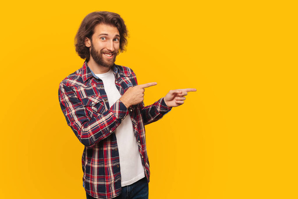 Cheerful young man with curly hair in a shirt points aside to free space, on a yellow background - Foto, afbeelding