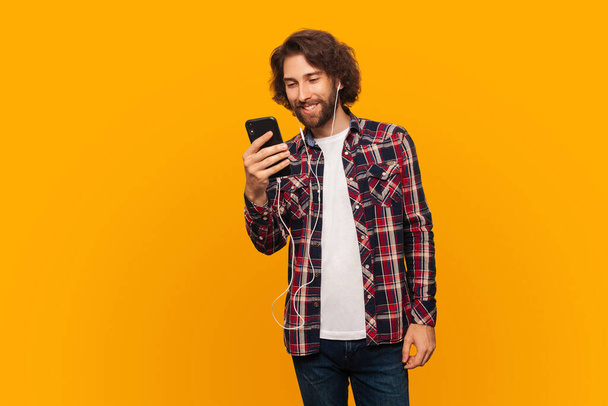 Šťastný mladý muž s kudrnatými vlasy v košili poslouchá hudbu na sluchátkách a drží smartphone. Muž ve sluchátkách drží mobilní telefon - Fotografie, Obrázek