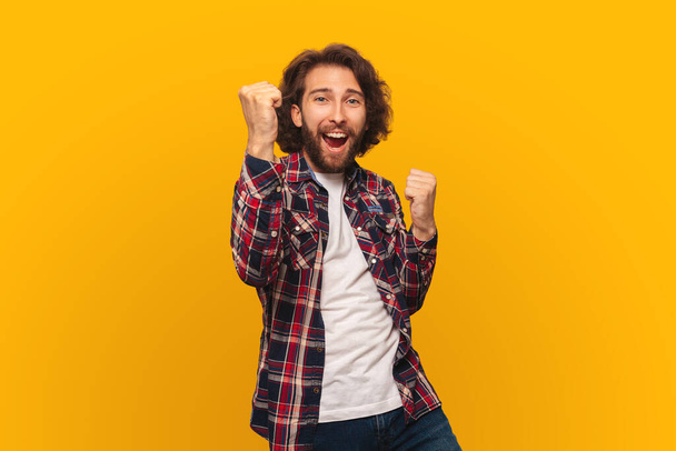Happy young man with long hair in a shirt celebrates victory with arms up on a bright yellow background - Фото, зображення