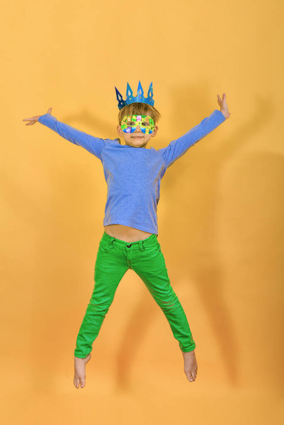 A cheerful and joyful boy in a paper crown is jumping on a yellow background in the studio. - Photo, Image