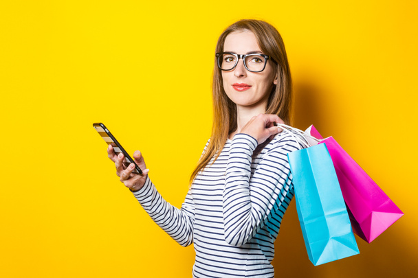 Friendly young woman holds shopping bags and phone on a yellow background - Foto, Bild