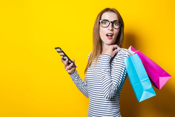 Giovane donna sorpresa con borse della spesa e con un telefono su uno sfondo giallo - Foto, immagini