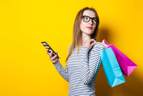 Mujer joven mira hacia atrás, sostiene un teléfono y bolsas de compras sobre un fondo amarillo
 - Foto, Imagen