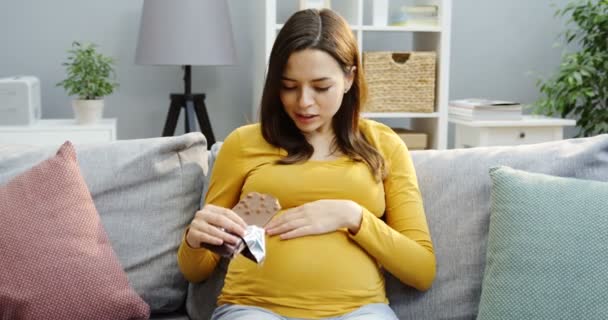 Beautiful pregnant woman eating a chocolate bar with nuts while sitting on the gray sofa with pillows in the cozy modern living room. Inside - Video