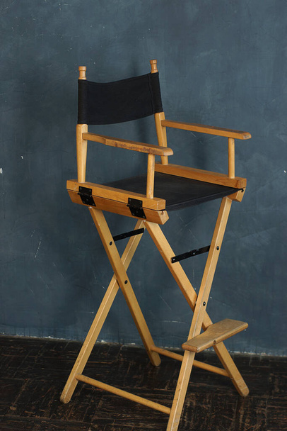 A filmmakers chair with wooden legs and a black seat stands against a gray-blue wall. - Foto, Imagen
