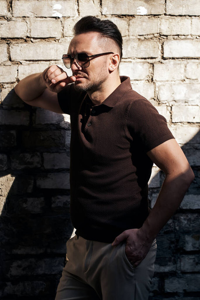 stylish guy with glasses stands near a stone wall and looks away. thought. strong and strict male gaze. brown T-shirt with collar and fashionable beige trousers. sunlight. half-length portrait. - Foto, afbeelding