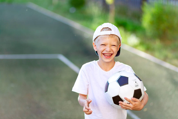 ritratto di un ragazzo biondo ridente con un berretto in uniforme sportiva con un pallone da calcio tra le mani sul campo di calcio. - Foto, immagini