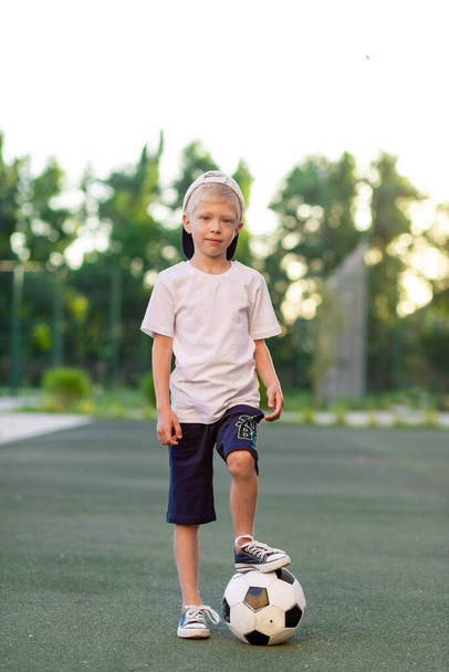 6-летний блондин стоит на зеленой лужайке на футбольном поле с футбольным мячом
. - Фото, изображение