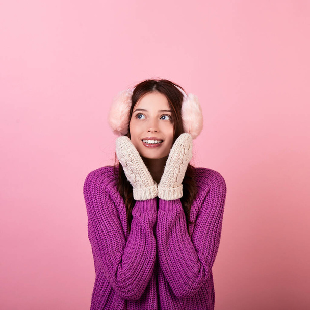 Cute young teenager in a knitted sweater and mittens and fluffy winter pink headphones. The girl smiles gently looks to the side and presses her hands to her face. Photo on pink background.Copy space. - Fotó, kép