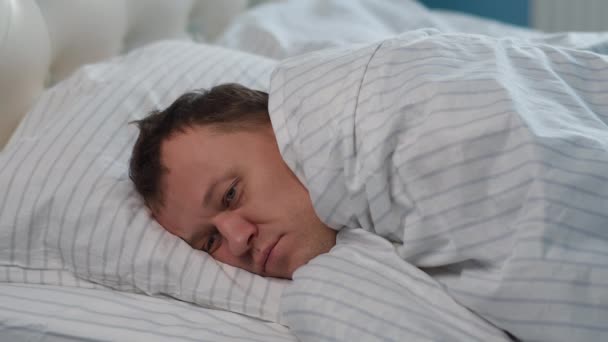 Een jonge man ligt op het bed bedekt met een deken, kan niet slapen - Video