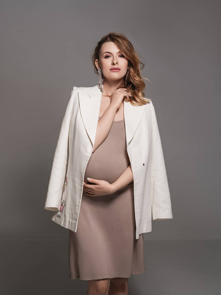 Eine junge schwangere Frau in einem schönen Kleid und einer weißen Jacke posiert im Studio.Schöne schwangere Frau berührt ihren Bauch mit den Händen auf einem grauen Hintergrund. Junge Mutter Vorfreude auf das Baby.Schwangerschaftsmode - Foto, Bild