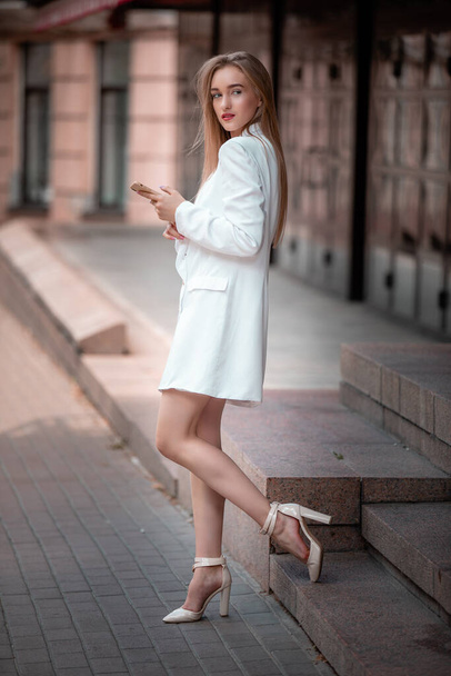 Portrait of Beautiful young business woman with phone at the hand on the summer street. Female model with long hair wearing at white suit standing at the city - Foto, immagini