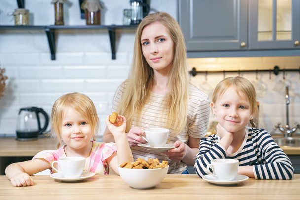 Portrait of a happy family young mother and two daughters drinking tea or milk with freshly baked homemade cookies in the kitchen. - Fotoğraf, Görsel