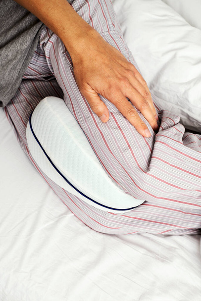 closeup ενός καυκάσιου άνδρα, φορώντας ριγέ παντελόνι πιτζάμας, χρησιμοποιώντας ένα ανατομικό μαξιλάρι ανάμεσα στα πόδια του, ενώ είναι ξαπλωμένος σε ένα κρεβάτι που με λευκά σεντόνια - Φωτογραφία, εικόνα